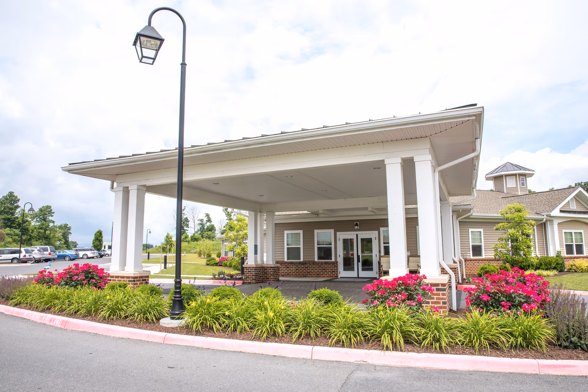 Entrance of Bellaire at Stone Port Assisted Living & Memory Care facility with a covered drop-off area supported by white columns, surrounded by landscaped greenery and bright pink flowers. A lamppost stands near the curb, and several cars are parked in the background under a partly cloudy sky.