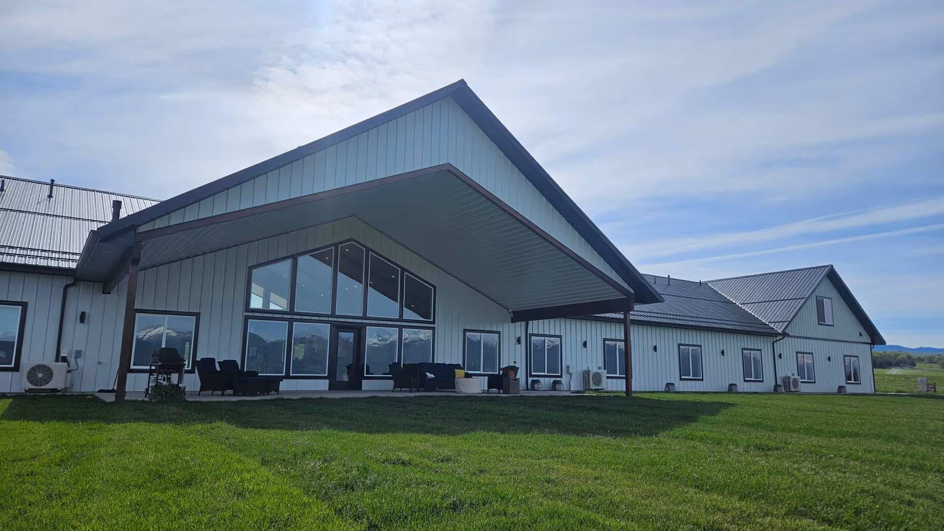 Exterior view of a single-story senior assisted living facility building with a large covered patio area furnished with outdoor seating. The building has white siding, multiple windows, and a metal roof. The surrounding area is a well-maintained green lawn under a partly cloudy sky.