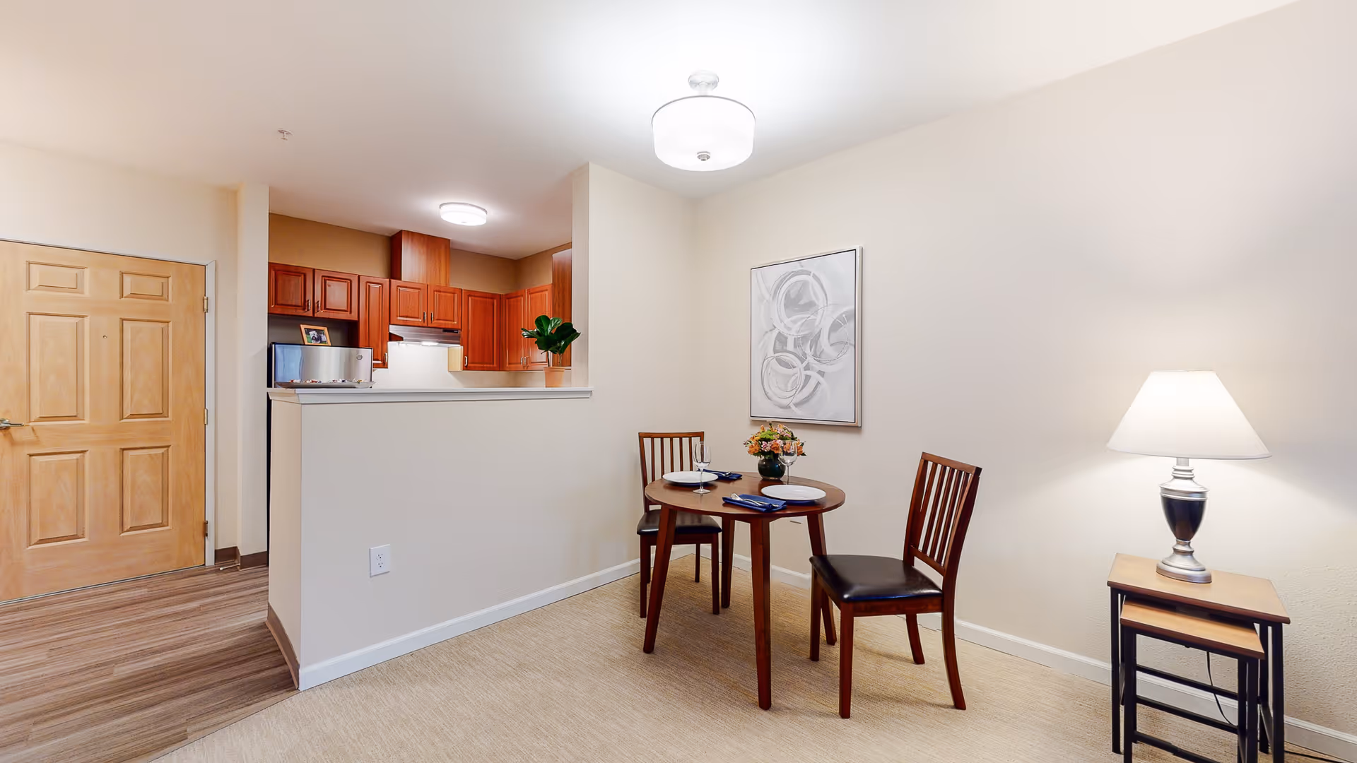 Dining area with a round table set for two, two chairs, a side lamp, and a view into a small kitchen and entry door.