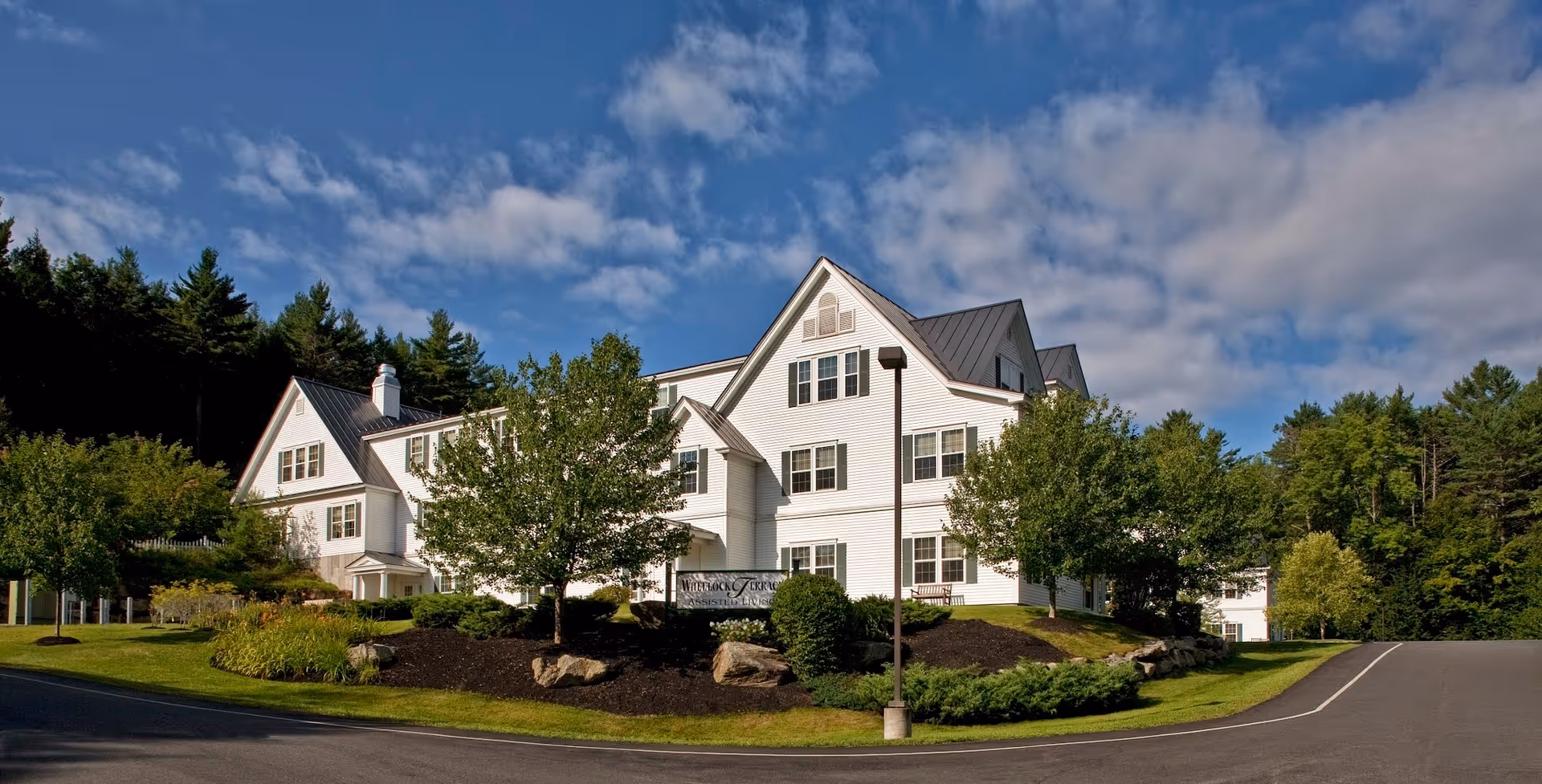 Exterior view of a large white multi-story building with a steep roof, surrounded by green trees and landscaping under a partly cloudy blue sky.