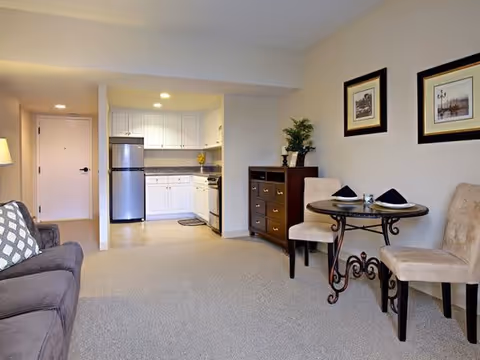 Open living area with a sofa, small round dining table and chairs, dresser, and a kitchenette with a stainless steel refrigerator.