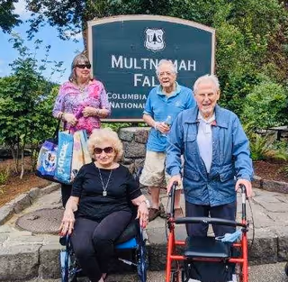 Four elderly individuals posing outdoors in front of a large sign that reads 'Multnomah Falls Columbia River National Scenic Area.' Two women and two men are present, one woman is seated in a wheelchair, and one man is using a walker. They are surrounded by greenery and trees.