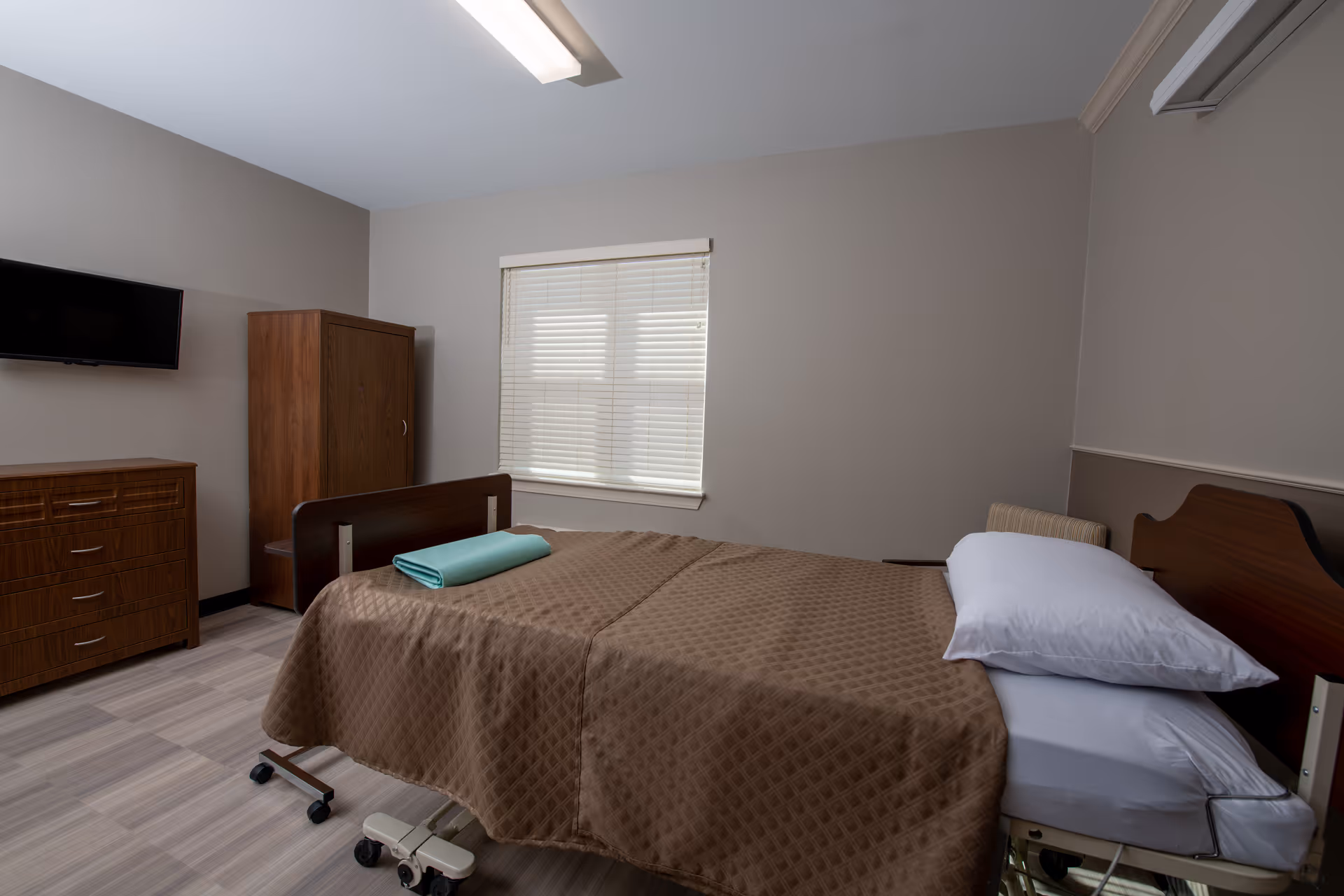 A simple, clean bedroom in a healthcare facility with a single hospital bed covered in a brown quilted blanket and a folded teal towel on top. The room has a wooden dresser, a wooden wardrobe, a wall-mounted flat-screen TV, and a window with closed blinds. The walls are painted light gray and the floor has light-colored tiles.