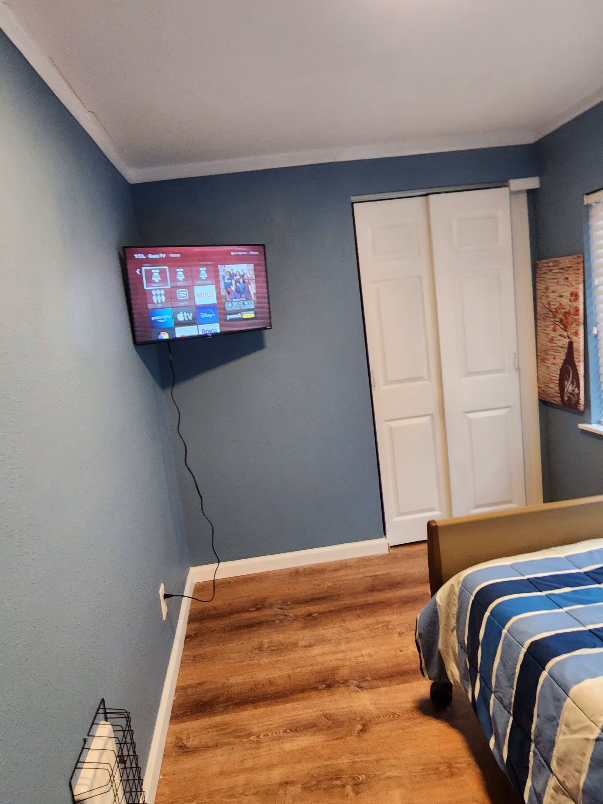 A bedroom with blue walls and wood flooring. A bed with a blue and white striped comforter is partially visible on the right side. A flat-screen TV is mounted on the left wall with a visible power cord plugged into an outlet near the floor. There is a white double-door closet in the center background and a window with blinds and a decorative wall hanging on the right.