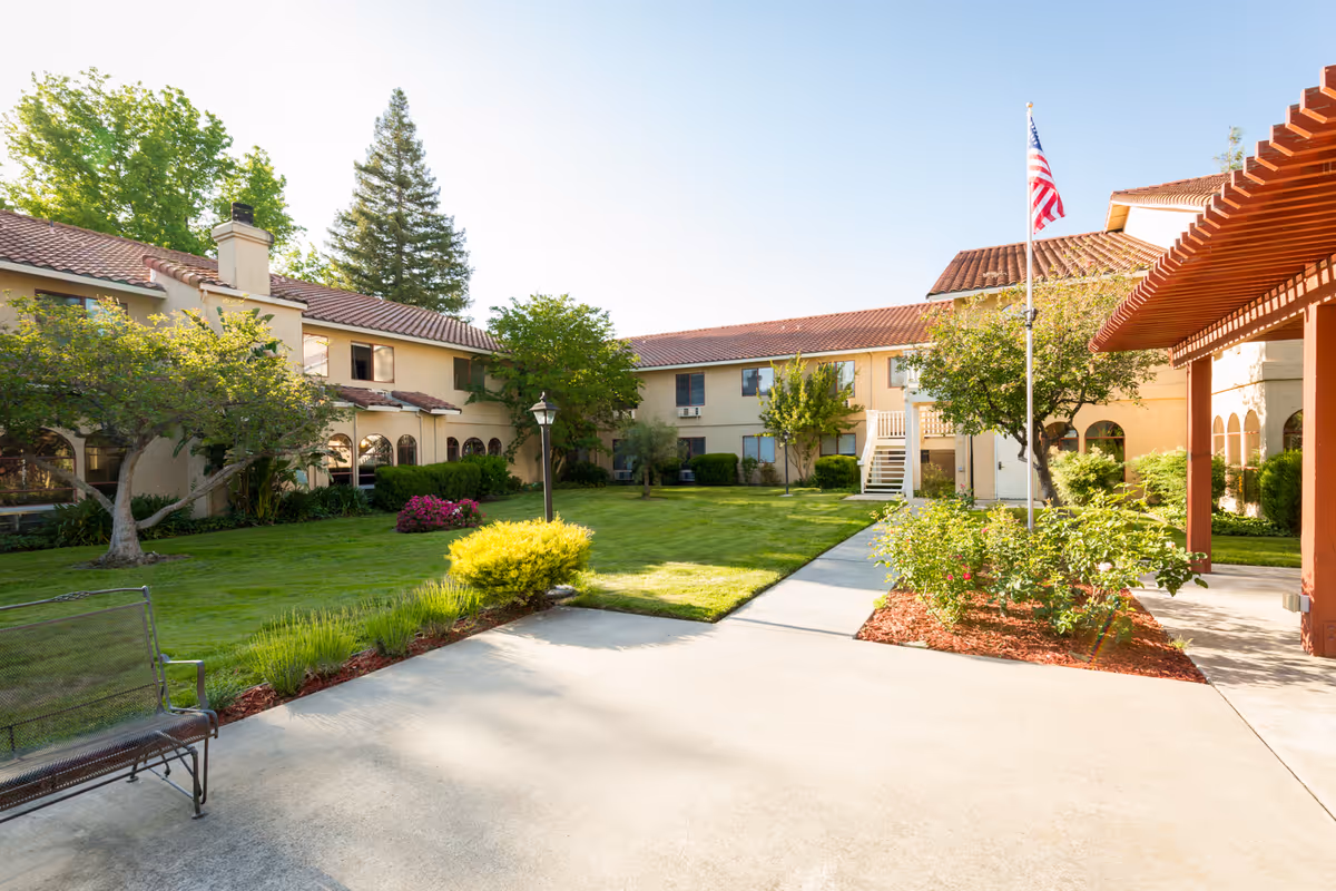Sunny landscaped courtyard with lawn, shrubs, a bench, an American flag, and a surrounding two-story senior living building.