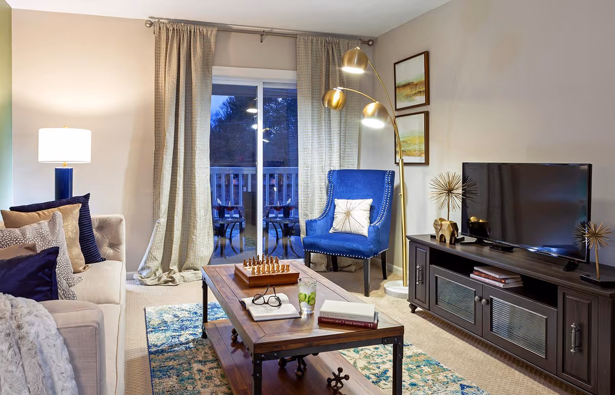 Well-decorated living room with a beige sofa, blue armchair, wooden coffee table and TV console in front of a sliding glass door with curtains.