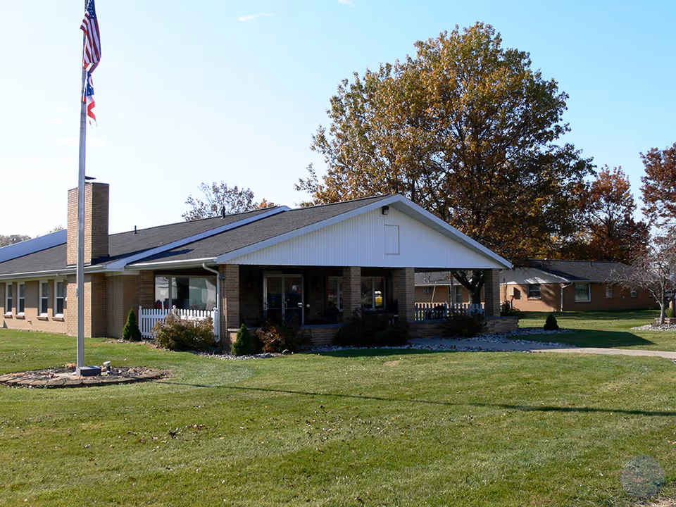 Exterior view of Oak Grove Healthcare Center, a single-story brick building with a covered entrance, surrounded by green lawn and trees with autumn foliage. An American flag and another flag are flying on a flagpole in front of the building.