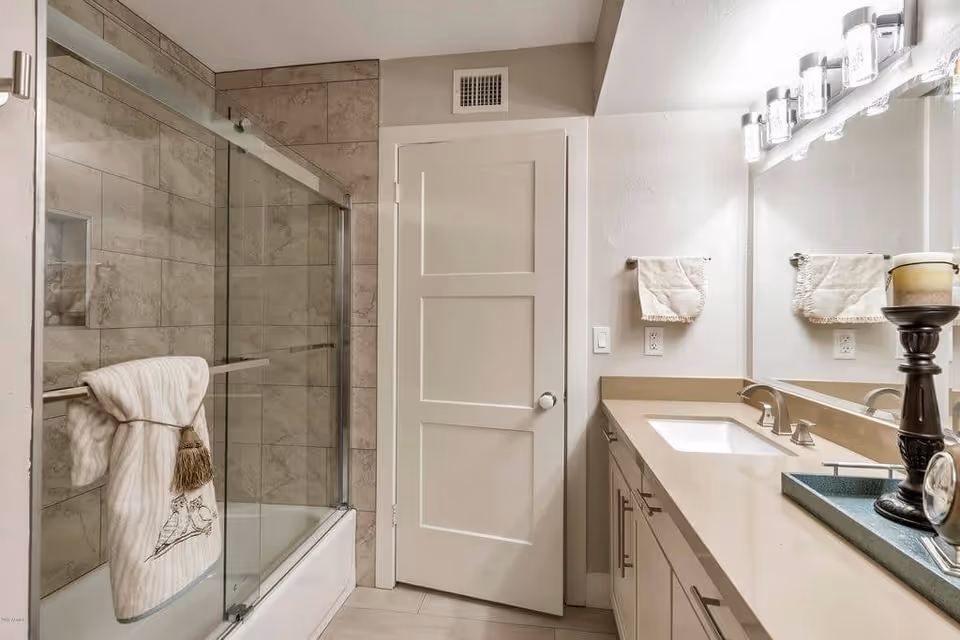 A modern bathroom with a glass-enclosed bathtub and shower on the left, beige tiled walls, a white door in the center, and a long countertop with a sink, faucet, and decorative items on the right. Two towels hang on towel bars, one on the shower door and one on the wall next to the mirror.