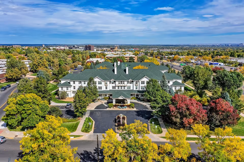 Aerial view of Woodlake Legacy Senior Living facility surrounded by trees with green, yellow, and red foliage. The building has a green roof and white exterior walls. The facility is located in a suburban area with roads, sidewalks, and other buildings visible in the background under a partly cloudy blue sky.