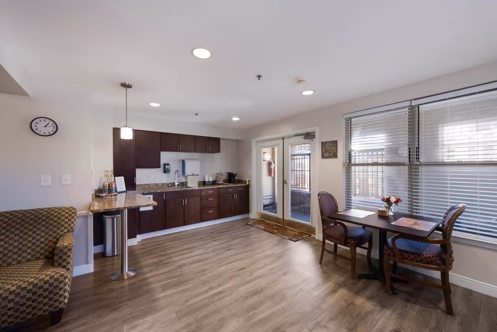 A bright and clean interior space featuring a small kitchenette with dark wood cabinets, a granite countertop, and a sink. To the right, there is a small dining area with a table and two chairs next to large windows with blinds. A double glass door leads to an outdoor patio area. On the left side, there is a patterned armchair and a wall clock showing the time as 1:10.