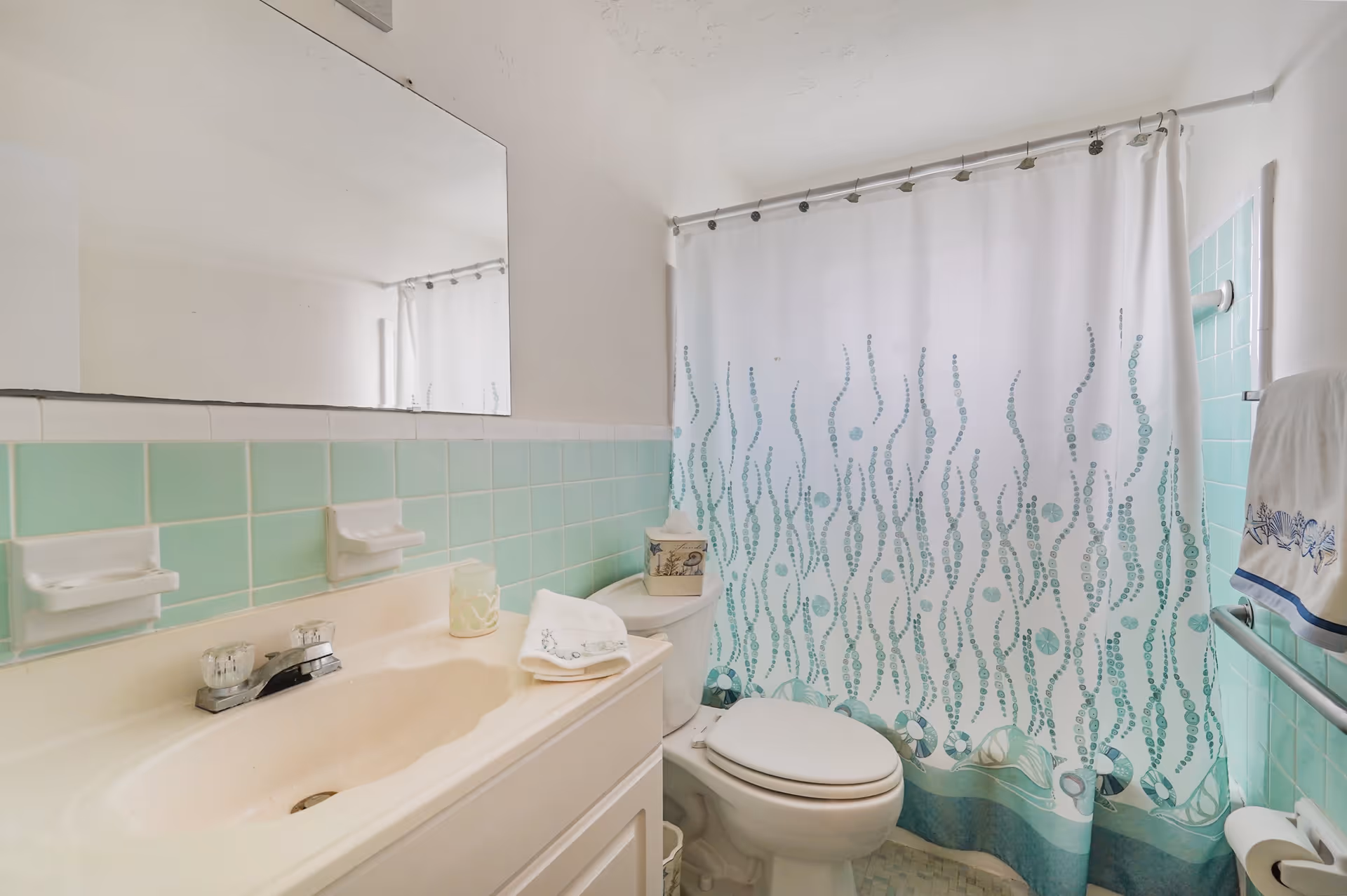 Small mint-green tiled bathroom with a sink, toilet, and a patterned shower curtain.