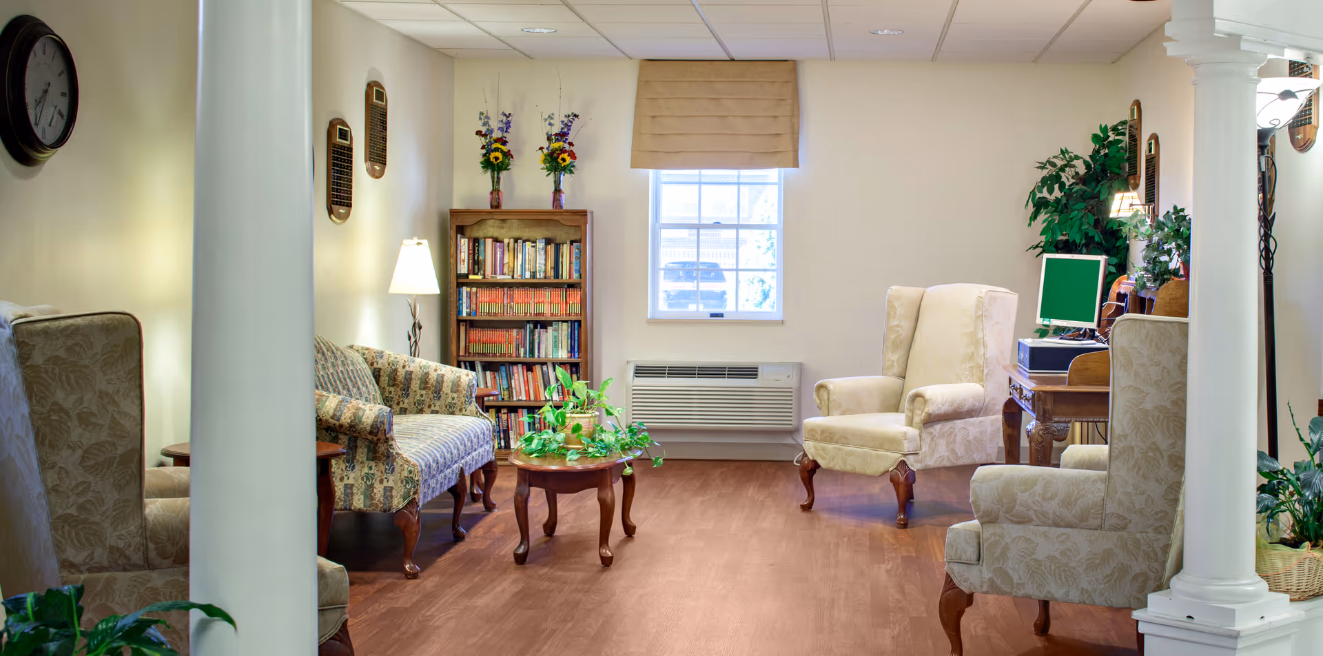 A cozy living room area in Hampshire Center featuring upholstered armchairs and a loveseat arranged around a wooden coffee table with a green plant. There is a bookshelf filled with books and two vases with flowers on top. A window with a beige valance lets in natural light. A computer desk with a monitor and keyboard is visible on the right side, along with several potted plants. The room has wooden flooring and white walls with decorative plaques and a clock.