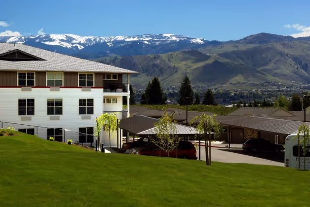 View of a senior living facility building with a green lawn in the foreground and covered parking spaces. Snow-capped mountains and a clear blue sky are visible in the background.