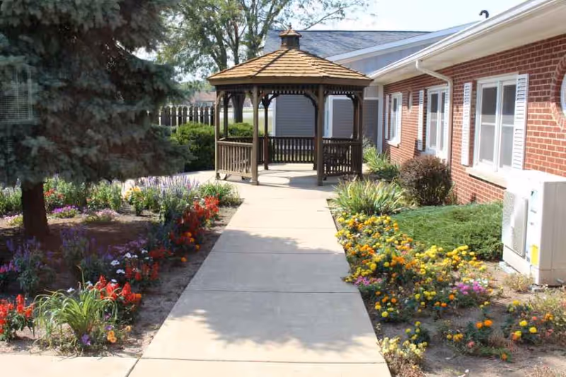 A paved walkway leading to a wooden gazebo surrounded by colorful flower beds and greenery, with a brick building on the right side and a large evergreen tree on the left.