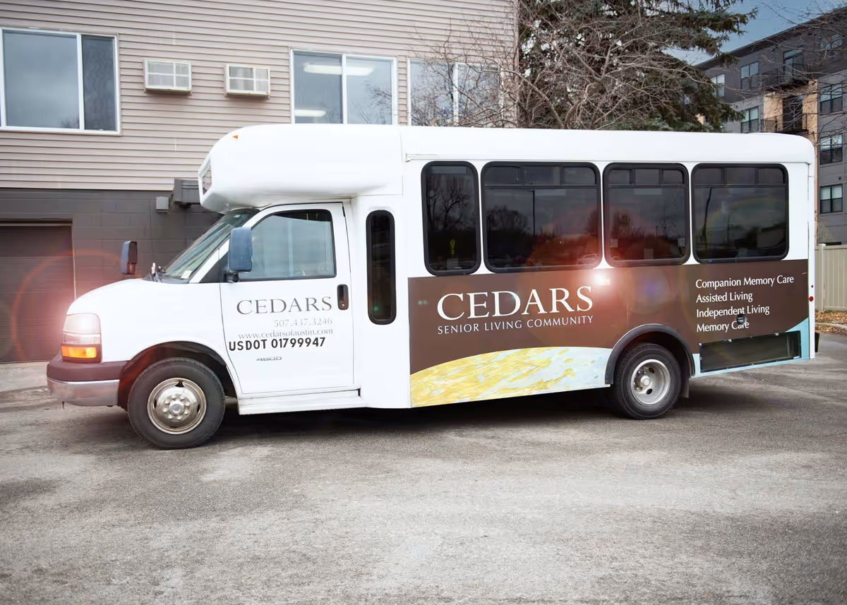 A white and brown shuttle bus parked outside a building. The bus has the text 'CEDARS SENIOR LIVING COMMUNITY' on the side along with services listed: Companion Memory Care, Assisted Living, Independent Living, Memory Care. The front door area displays contact information and a USDOT number.