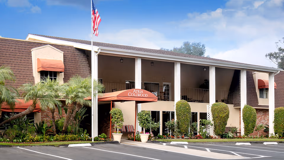 Exterior view of Atria Collwood senior living facility showing a two-story building with a covered entrance, an American flag on a flagpole, well-maintained landscaping with palm trees and shrubs, and a parking area in front.