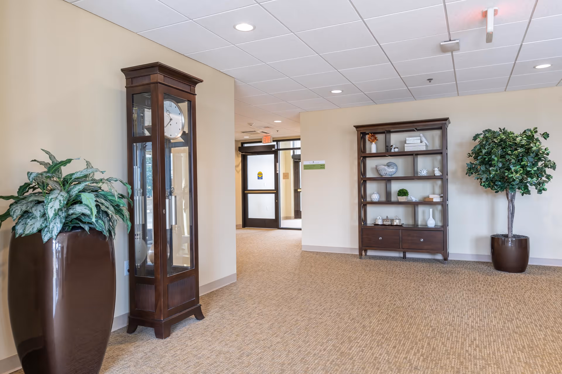 Bright interior lobby with a tall grandfather clock, decorative shelving, and large potted plants.