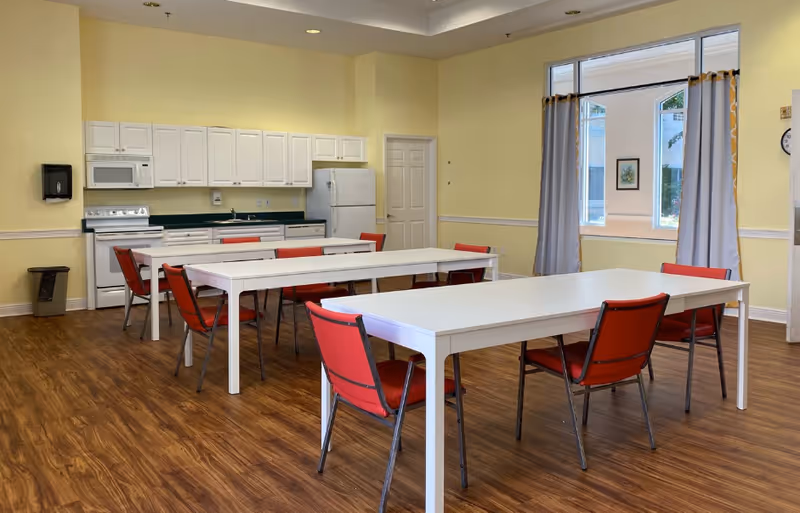 Communal dining room with white tables and red chairs, a kitchenette along the back wall, and a large window with curtains.