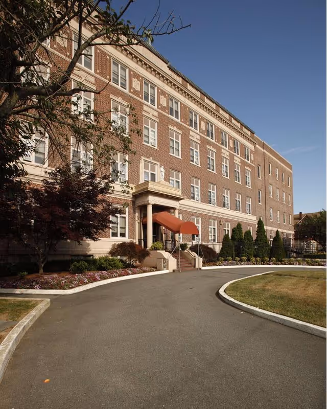 Exterior view of a multi-story brick building with white trim and many windows, featuring a red awning over the entrance. The building is surrounded by landscaped greenery, including trees, shrubs, and flower beds, with a curved driveway leading to the entrance under a clear blue sky.