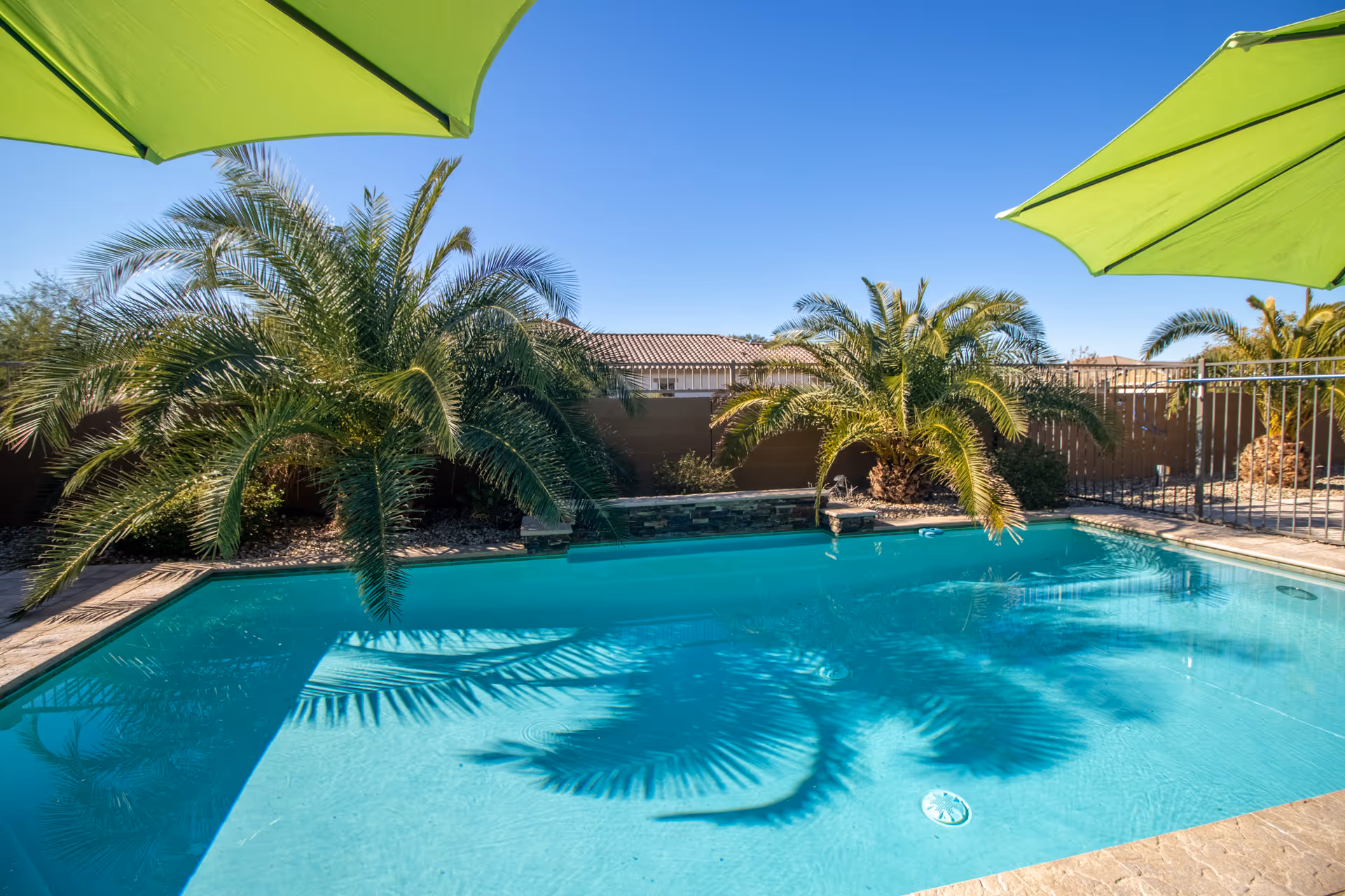 Outdoor swimming pool with clear blue water surrounded by palm trees and green umbrellas under a bright blue sky.