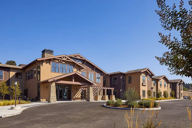 Exterior front facade of a two-story senior living facility with a covered entrance, stone columns, and landscaped driveway under a clear blue sky.