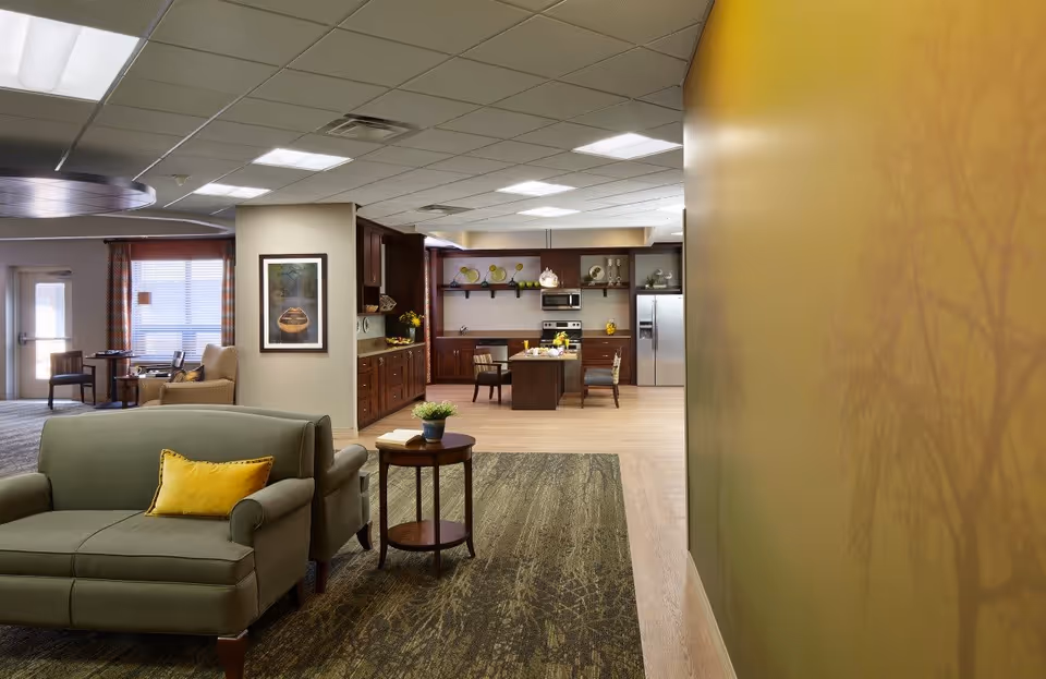 Interior view of a senior living facility showing a cozy seating area with a green loveseat and a yellow pillow, a small round wooden table with a plant and a book, and an open kitchen area in the background with wooden cabinets, a stainless steel refrigerator, stove, and a small dining table with chairs. The walls are painted in warm tones, and there is a large yellow wall with a tree shadow pattern on the right side.
