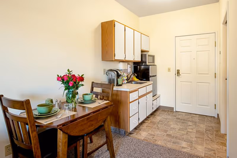 Small kitchenette and dining area with a set wooden table, cabinets, compact appliances, and an entry door.
