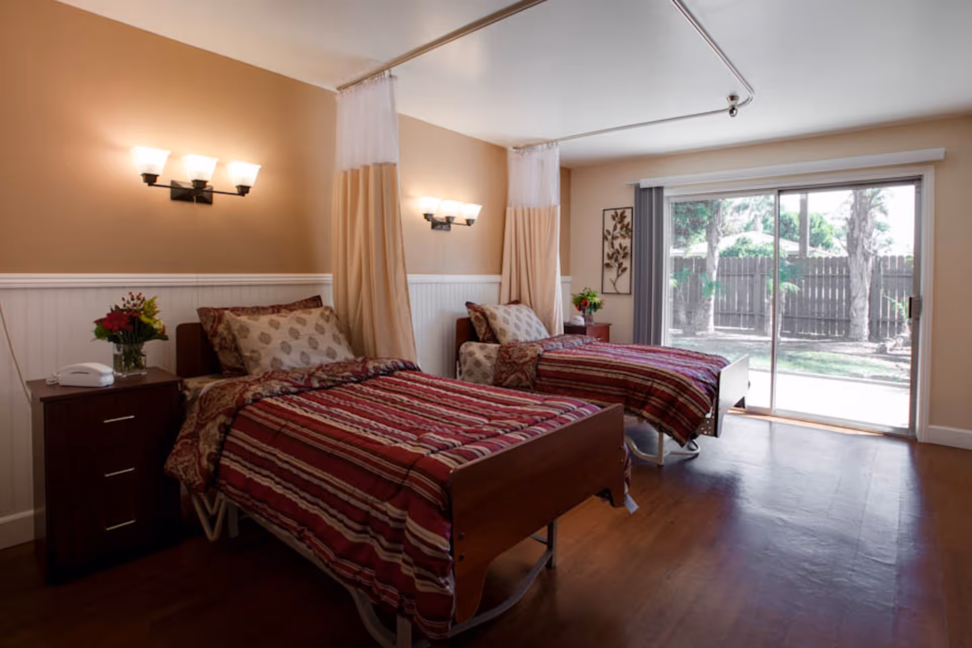 A room with two single beds separated by privacy curtains, each bed has a striped red and beige bedspread and patterned pillows. There are wall-mounted lights above each bed, a nightstand with a telephone and a vase of flowers, and a large sliding glass door that opens to an outdoor area with trees and a wooden fence.