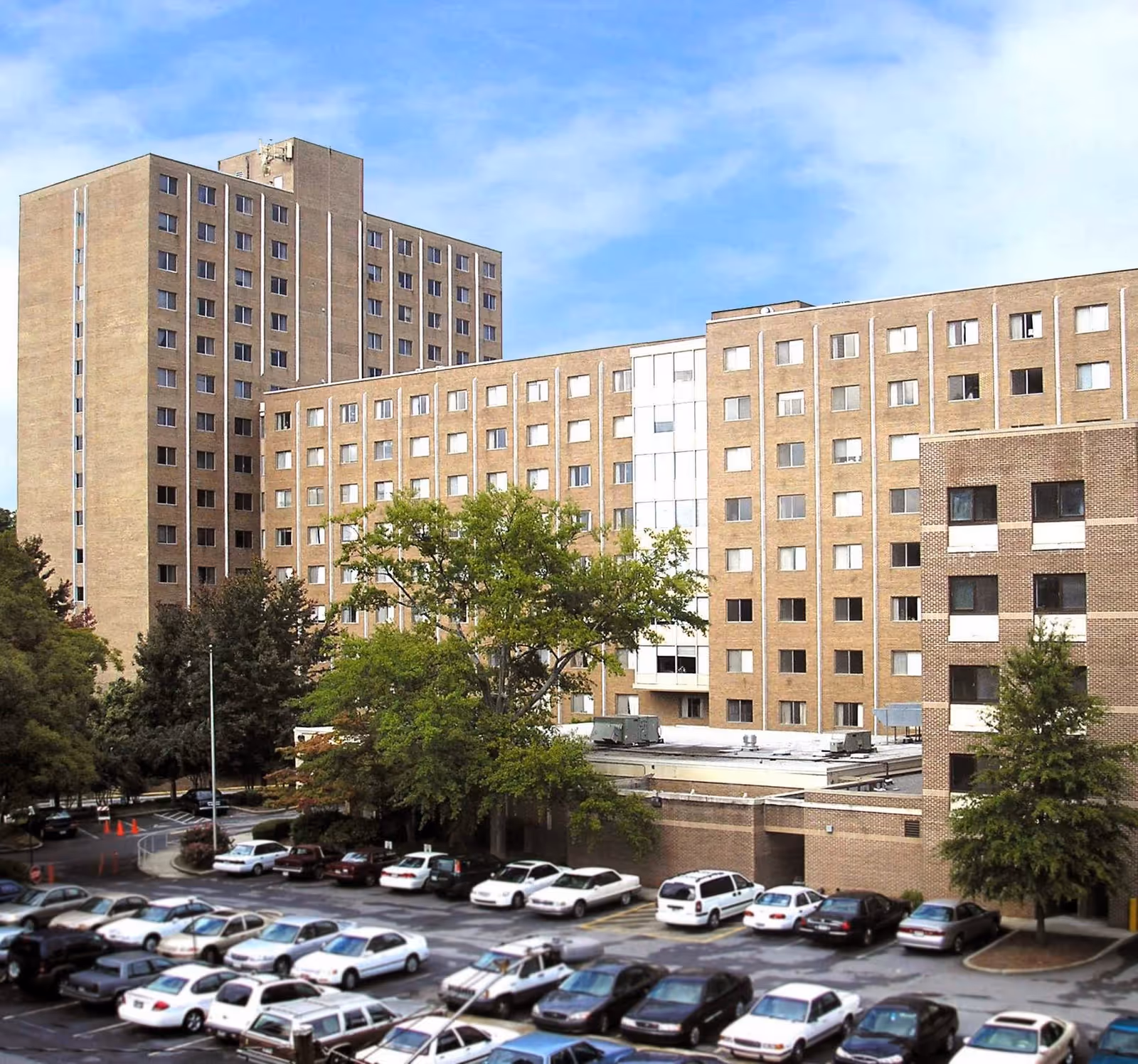 Exterior view of a large multi-story brick senior living building with a parking lot and trees in front.