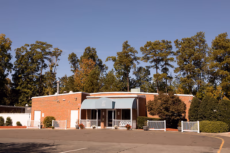 Exterior view of a single-story brick building with a light blue awning over the entrance. The building is surrounded by trees and bushes, with a clear blue sky overhead. There is a paved parking area in front of the building.