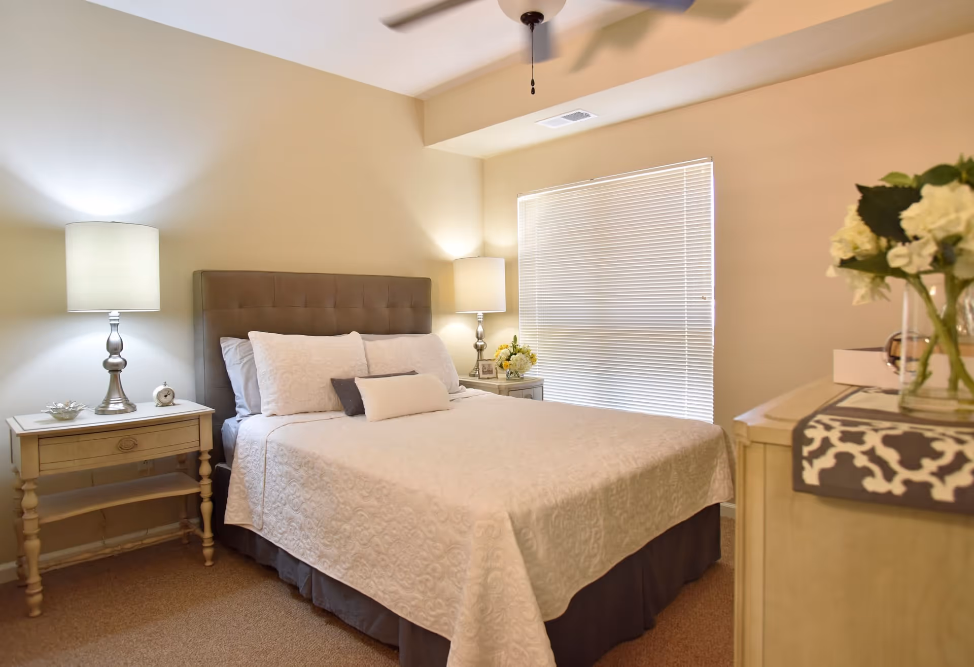 A cozy bedroom with a neatly made bed featuring white bedding and multiple pillows. There are two bedside tables, each with a lamp, and a window with closed blinds. A ceiling fan is visible above, and a dresser with a vase of flowers is partially visible on the right side.