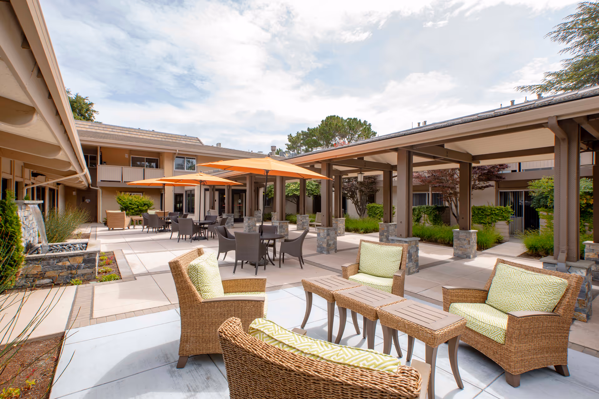 Outdoor courtyard area at BridgePoint at Los Altos featuring wicker chairs with green cushions, tables, and several orange umbrellas providing shade. The courtyard is surrounded by a two-story building with balconies and greenery, under a partly cloudy sky.