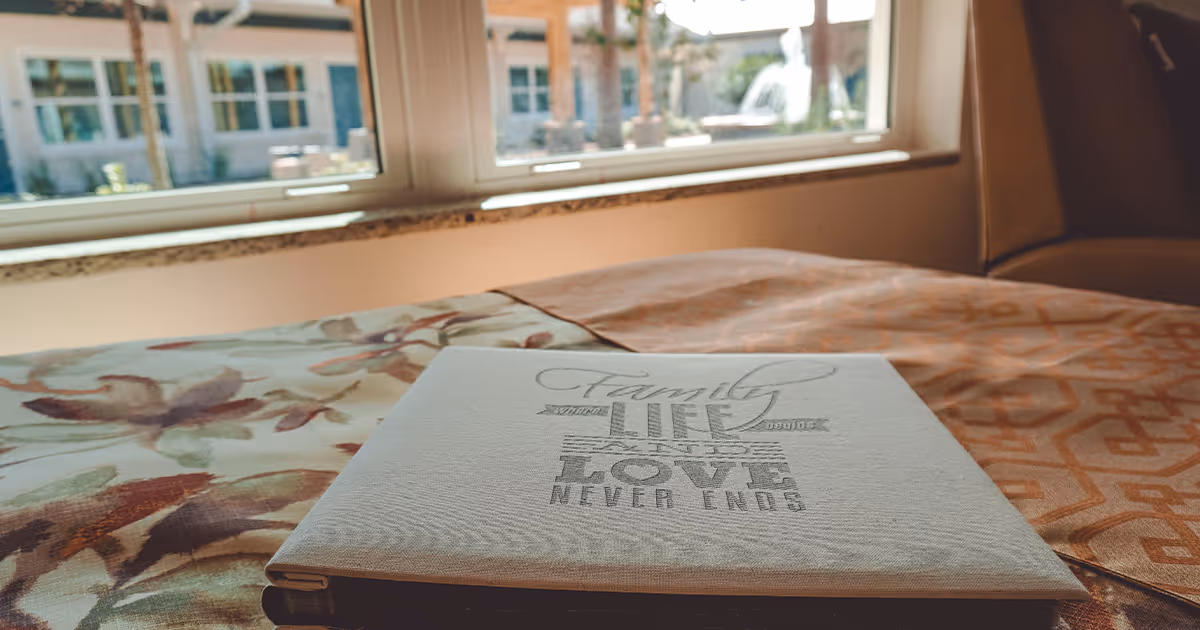 A close-up view of a bed with a floral and patterned bedspread, a book or album with the words 'Family where life begins and love never ends' on the cover, and a window showing an outdoor courtyard with a fountain and building in the background.