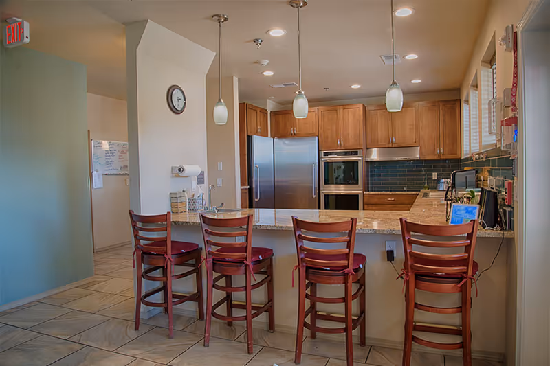 Interior view of a kitchen area with a granite countertop island and four wooden bar stools with red cushions. The kitchen features wooden cabinets, stainless steel refrigerator, built-in oven, and a green tiled backsplash. Three pendant lights hang above the island. A clock is mounted on a white wall, and an exit sign is visible above a doorway to the left.