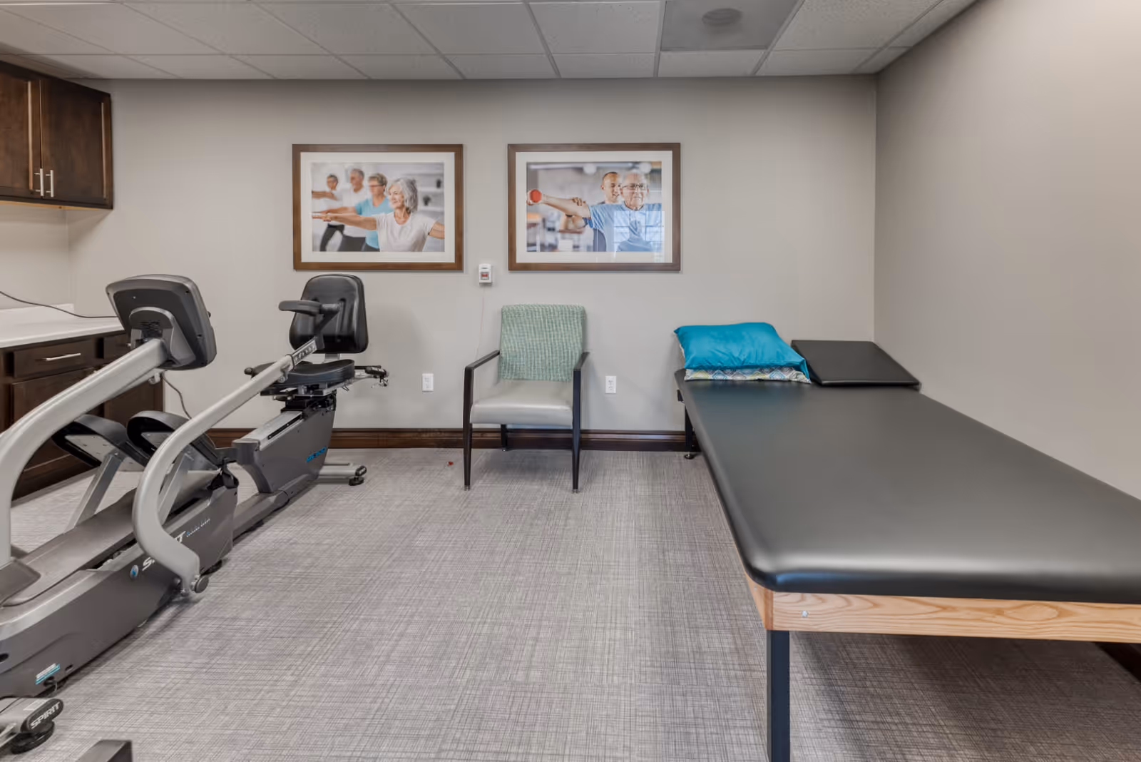 A small exercise room with a recumbent stationary bike, a padded examination or therapy table with pillows, a single chair with a green throw draped over the back, and two framed photos on the wall showing elderly people exercising.
