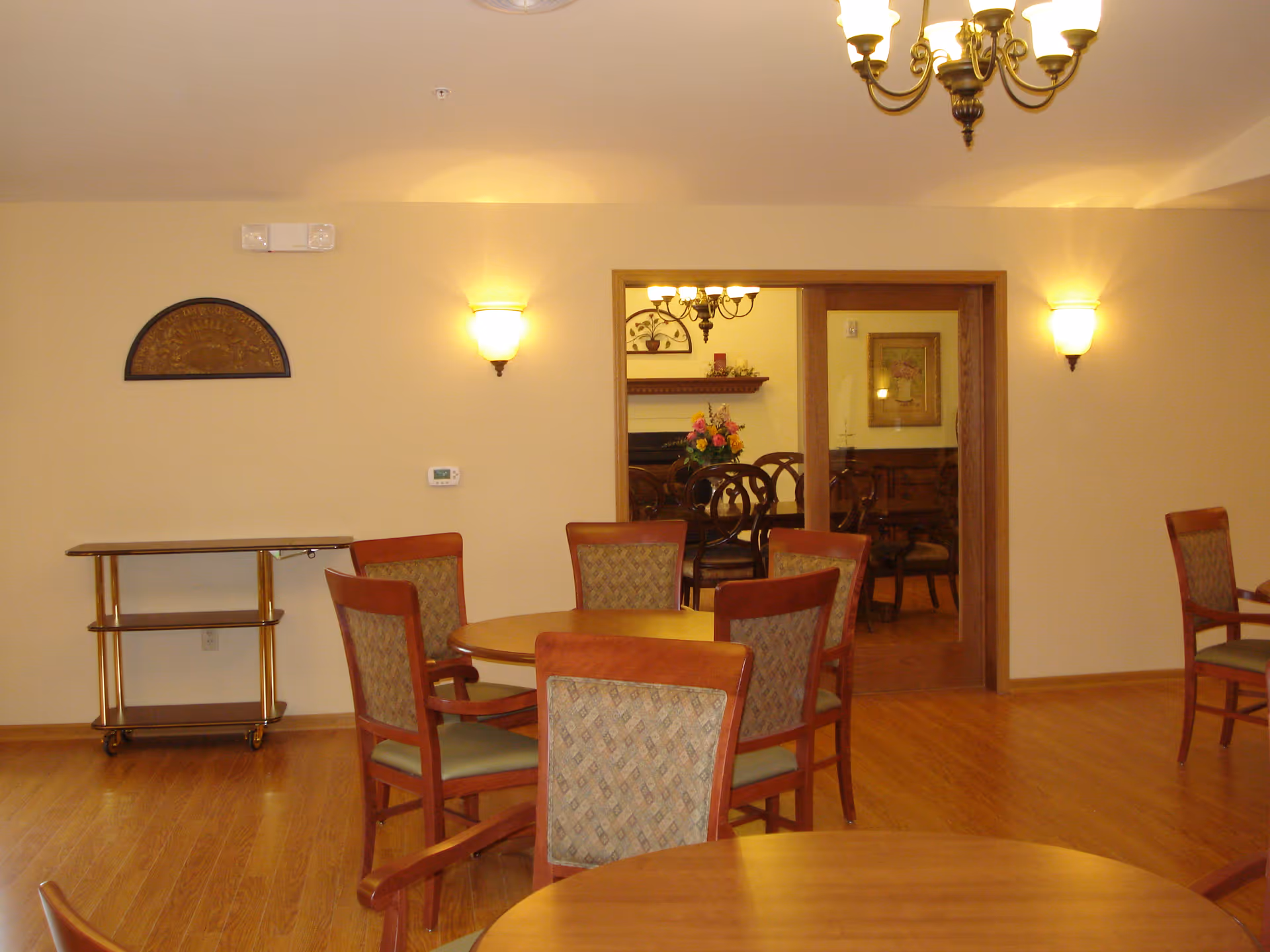 Interior view of a dining area with wooden round tables and cushioned chairs. The room has wooden flooring, beige walls, and wall-mounted light fixtures. A doorway with glass sliding doors leads to another room with additional dining furniture and a floral arrangement on a table.