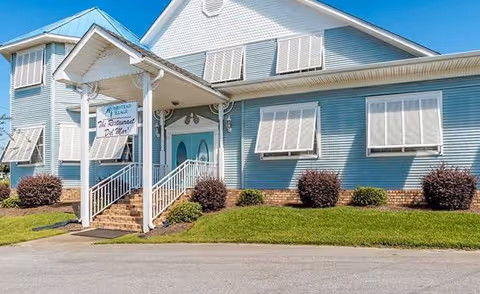 Exterior view of a light blue building with white trim and multiple windows with awnings. There is a covered entrance with stairs leading up to double doors. A sign near the entrance reads 'Homestead Village The Restaurant Del Mar'. The building is surrounded by neatly trimmed bushes and green grass under a clear blue sky.