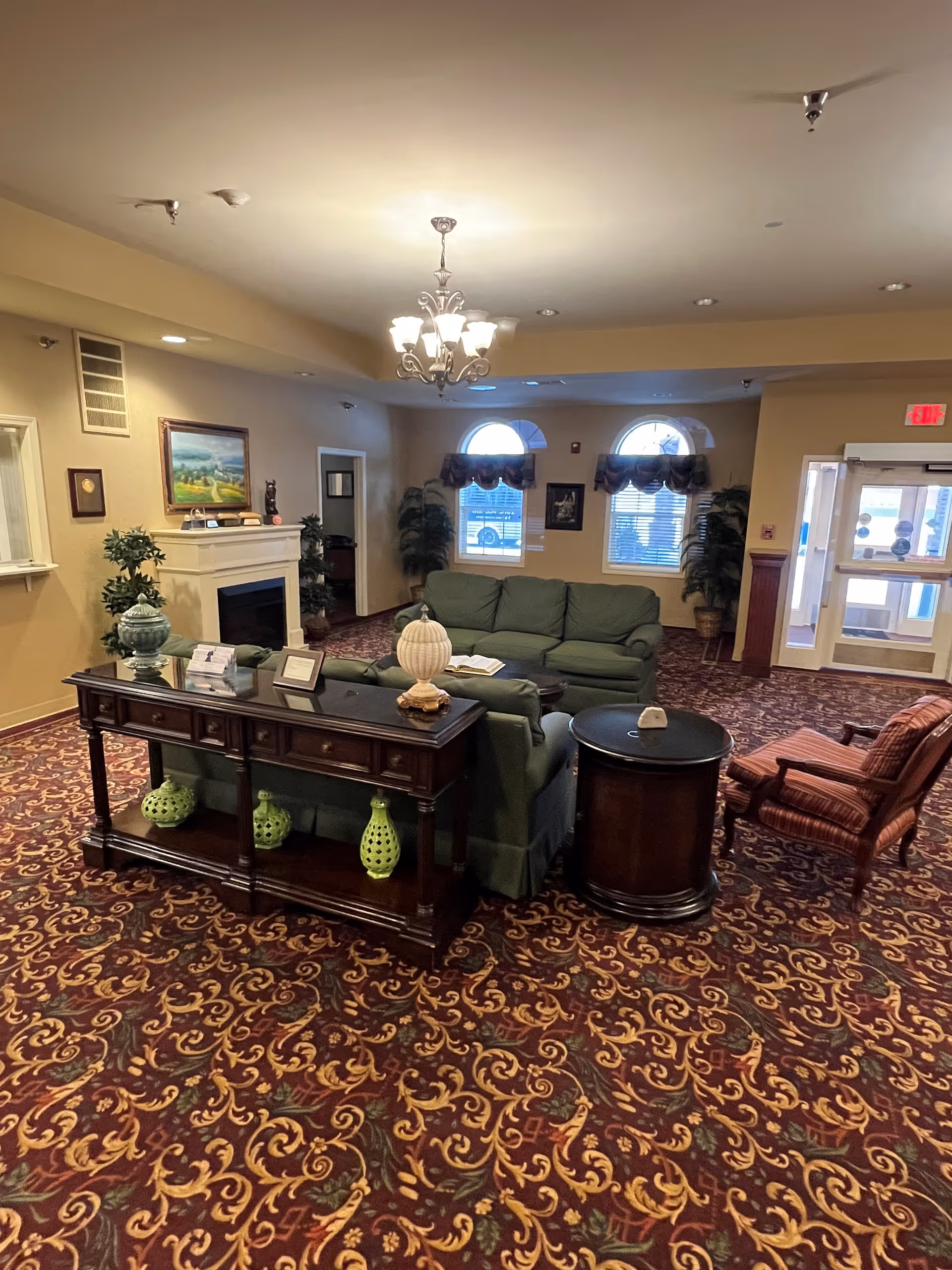 A seating lounge with green sofas, a wooden console table, fireplace, chandelier, and patterned carpet in a common area.