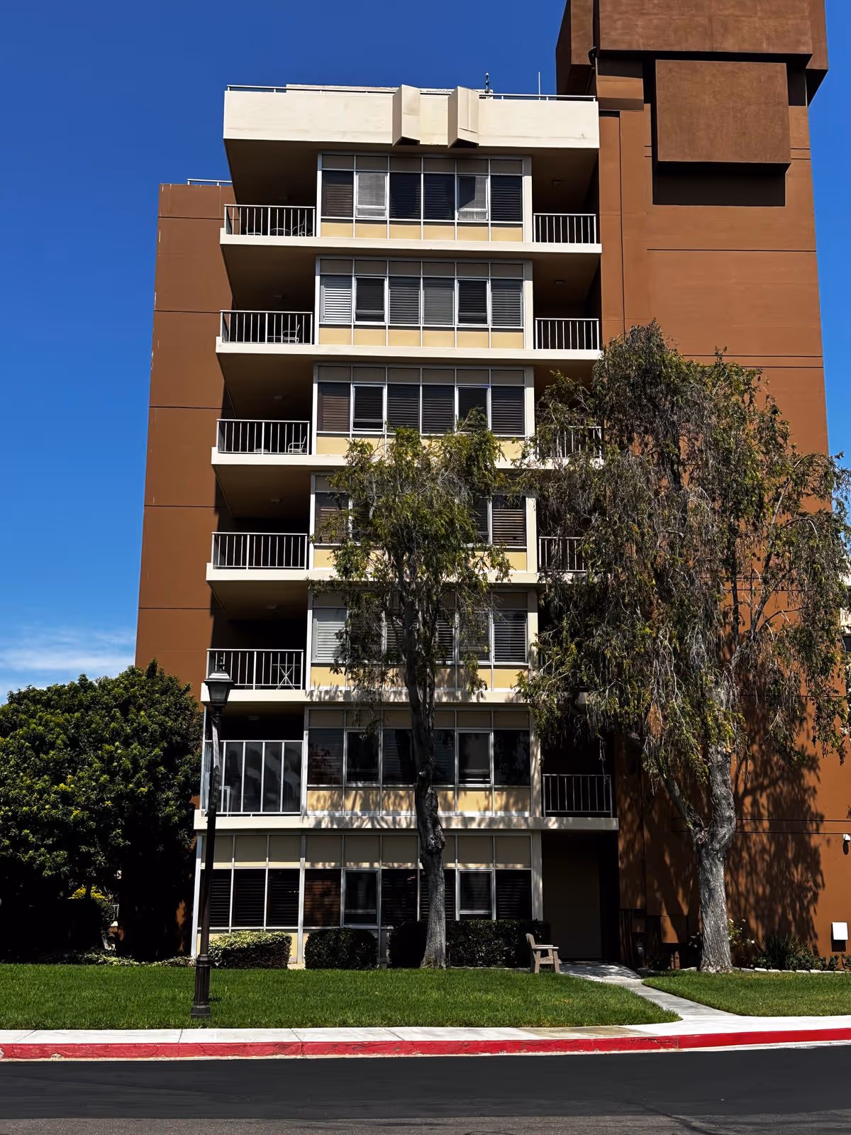 Front exterior of a multi-story brown apartment building with balconies, trees, and a grassy lawn.