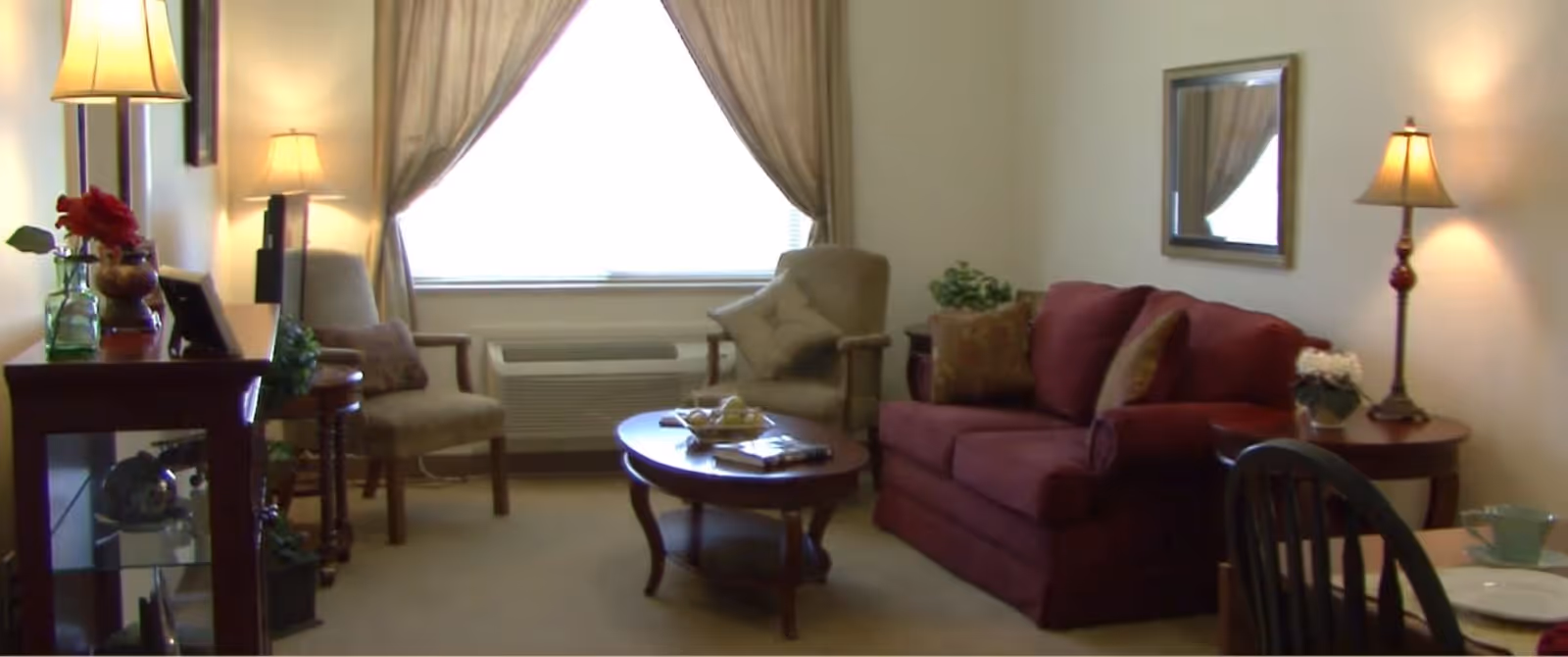 A cozy living room with a maroon sofa, two beige armchairs, a wooden coffee table with decorative items, a side table with a lamp and flowers, a mirror on the wall, and a large window with beige curtains letting in natural light.
