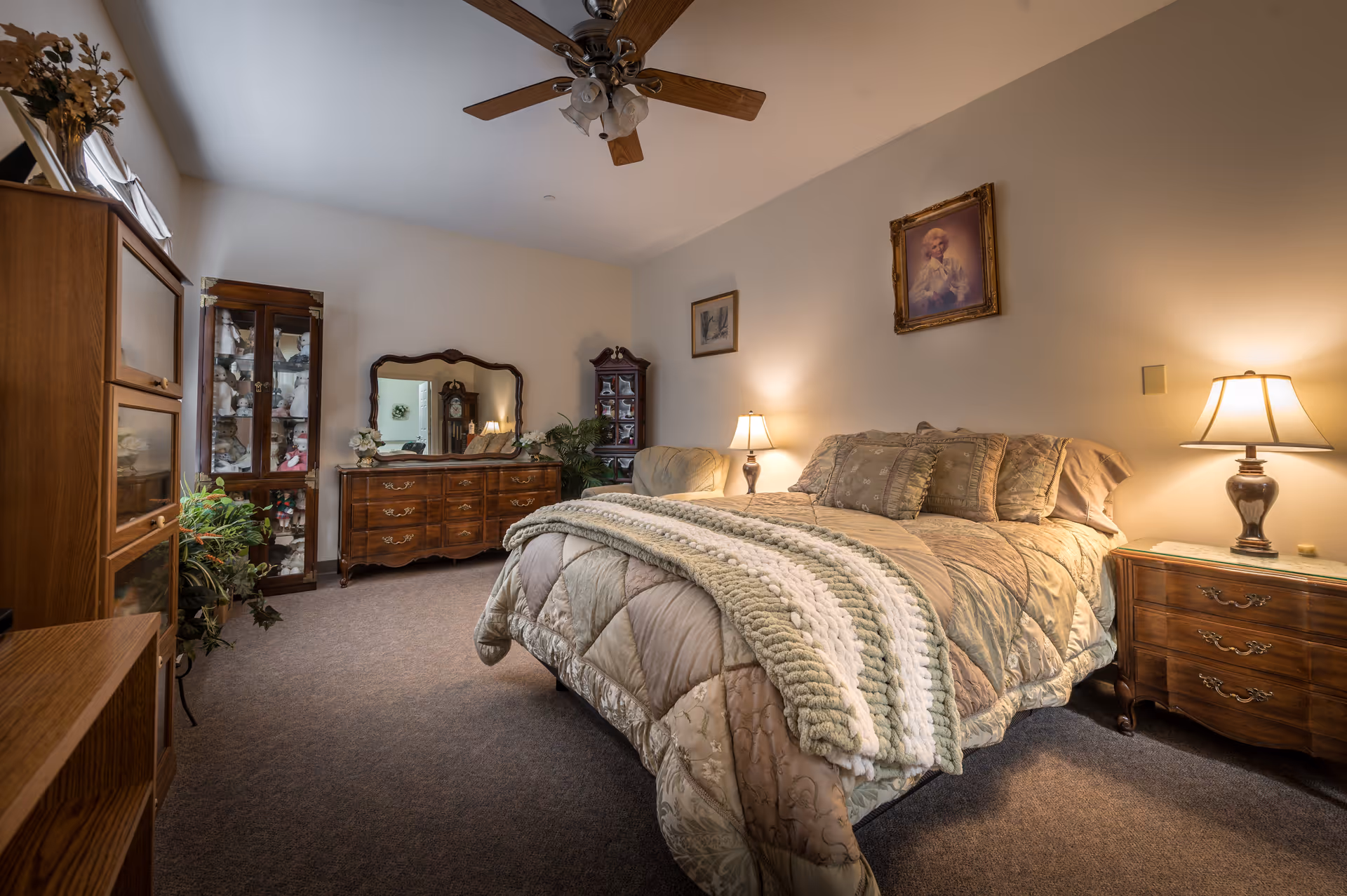 A cozy bedroom in an assisted living facility featuring a large bed with multiple pillows and a knitted throw blanket. The room has wooden furniture including a dresser with a mirror, a nightstand with a lamp, and a glass-front cabinet displaying decorative items. The walls are adorned with framed artwork, and a ceiling fan is mounted above.