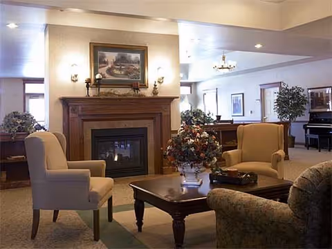 A cozy living room area in an assisted living facility featuring a fireplace with a painting above it, two beige armchairs, a floral centerpiece on a wooden coffee table, and additional seating with a piano visible in the background.