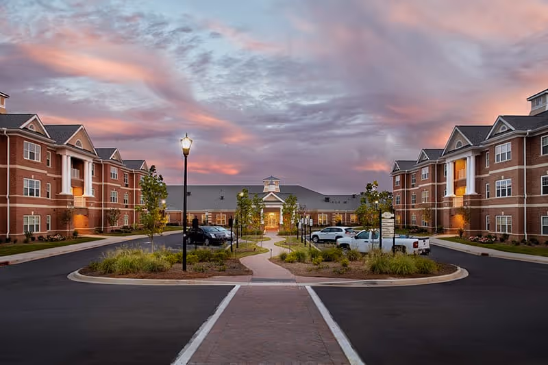 Exterior view of Wellmore of Lexington senior living facility at dusk, showing a paved driveway with parked cars, landscaped greenery, and two large brick buildings with white columns on either side under a colorful sky.