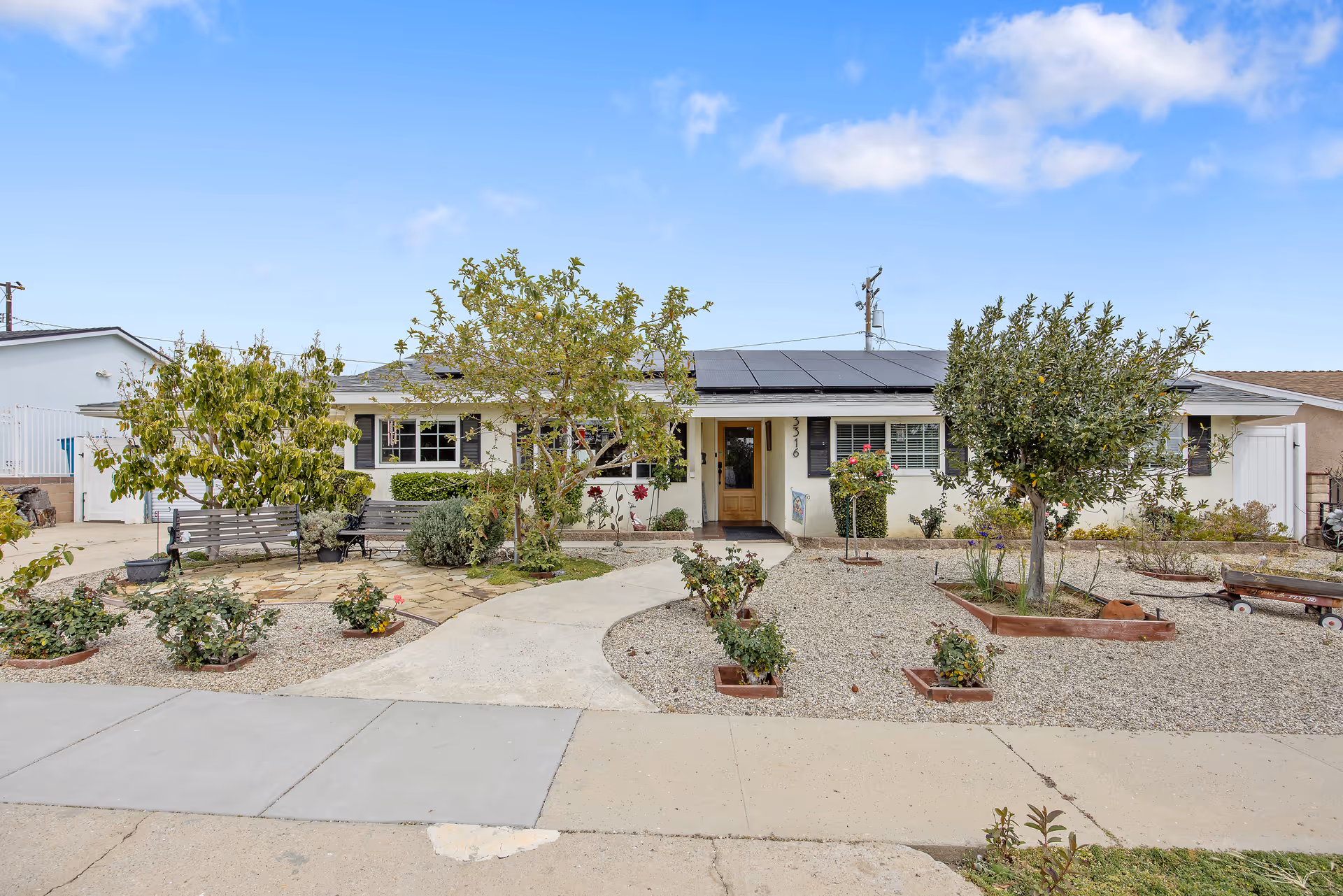 Single-story building with a front yard featuring trees, shrubs, and benches under a clear blue sky. The building has solar panels on the roof and a central entrance door with windows on either side.