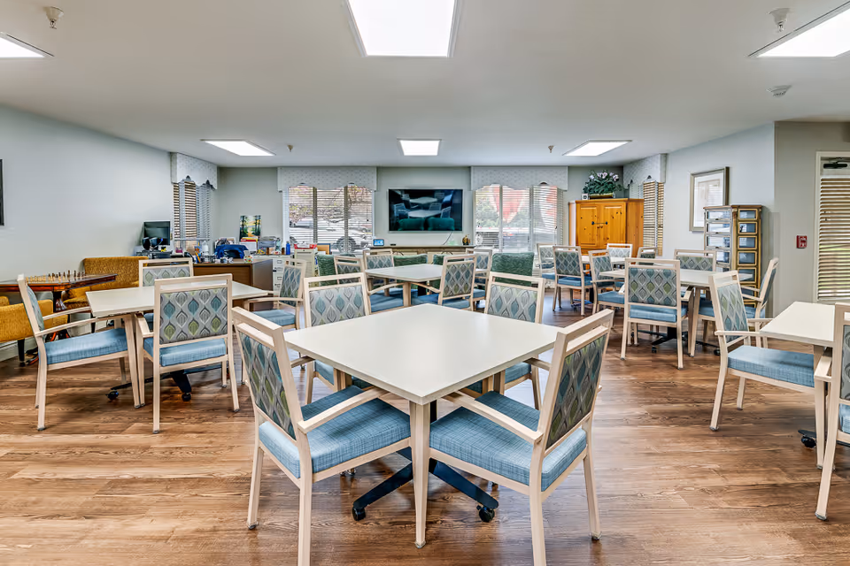 A bright and spacious common area with multiple square tables and cushioned chairs arranged around them. The room has large windows with blinds and valances, a flat-screen TV mounted on the wall, wooden flooring, and a mix of seating including armchairs and a bench. There are various decorative items and storage cabinets along the walls.