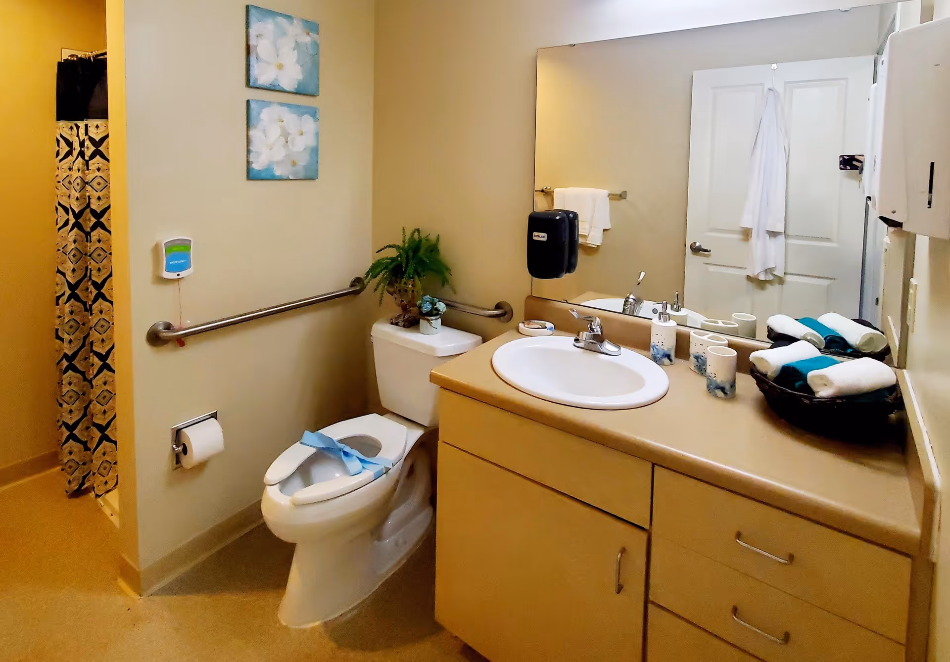 A clean and accessible bathroom with a toilet, sink, and countertop. The toilet has a blue ribbon on the seat, and there is a grab bar on the wall next to it. Above the toilet is a small plant and two floral paintings on the wall. The sink area has a large mirror, soap dispenser, cups, and a basket with rolled towels. A shower with a patterned curtain is visible in the background.