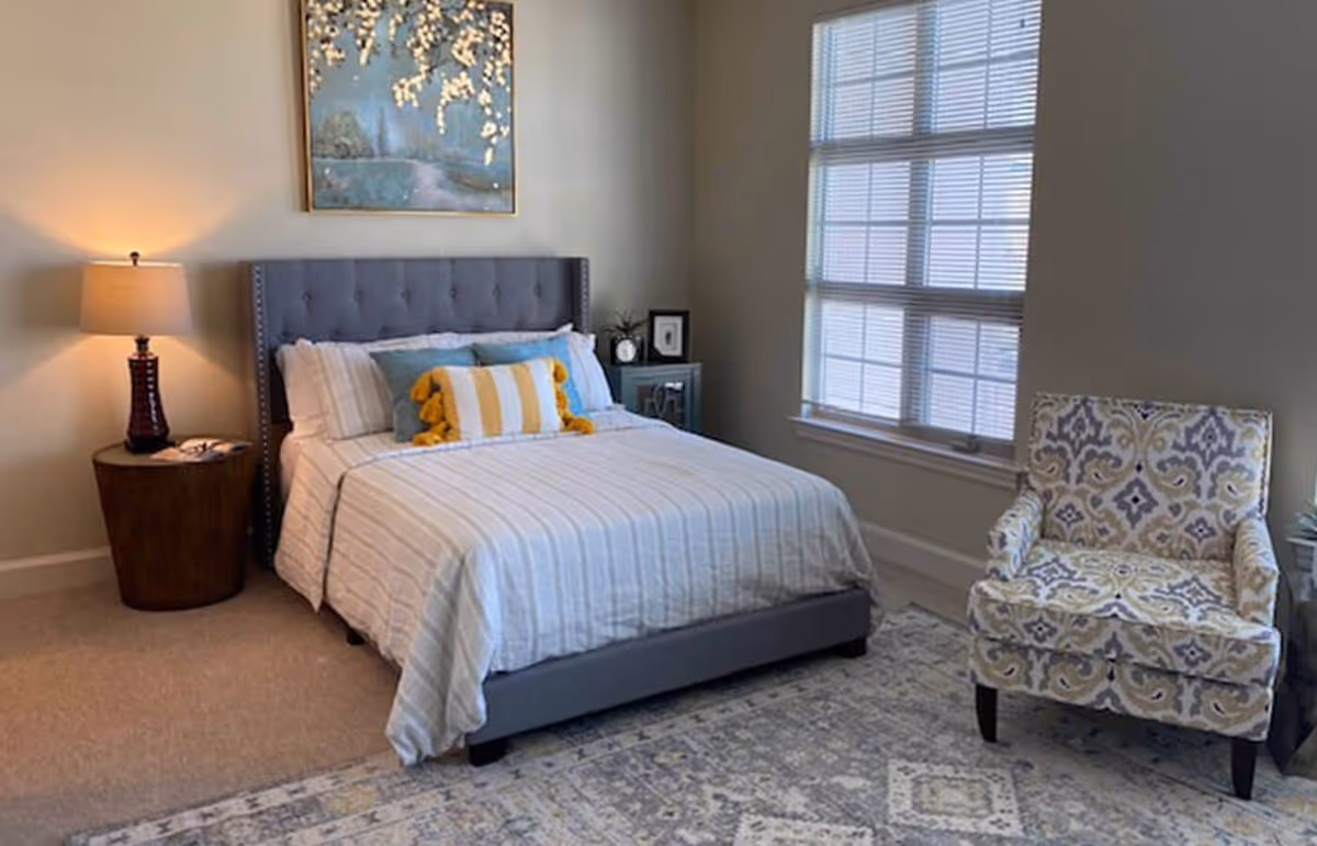 Neat bedroom with a gray upholstered bed, striped bedding, a bedside table and lamp, patterned armchair, large window, and wall art.