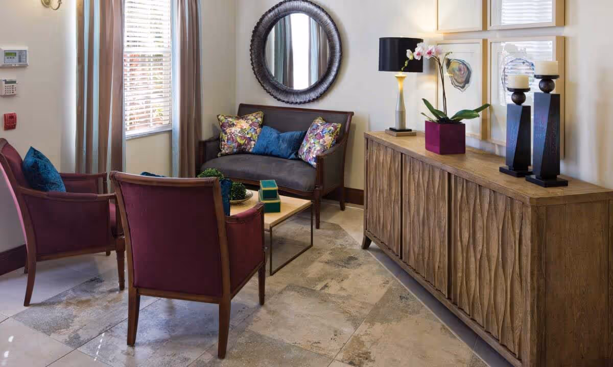 A cozy sitting area with two purple armchairs and a dark wooden loveseat adorned with colorful pillows. A small wooden coffee table sits in the center with decorative items. A wooden sideboard with a textured front holds a lamp, an orchid plant, and two candle holders. A round mirror hangs on the wall above the loveseat, and framed artwork is displayed above the sideboard. Light filters in through a window with blinds and curtains.