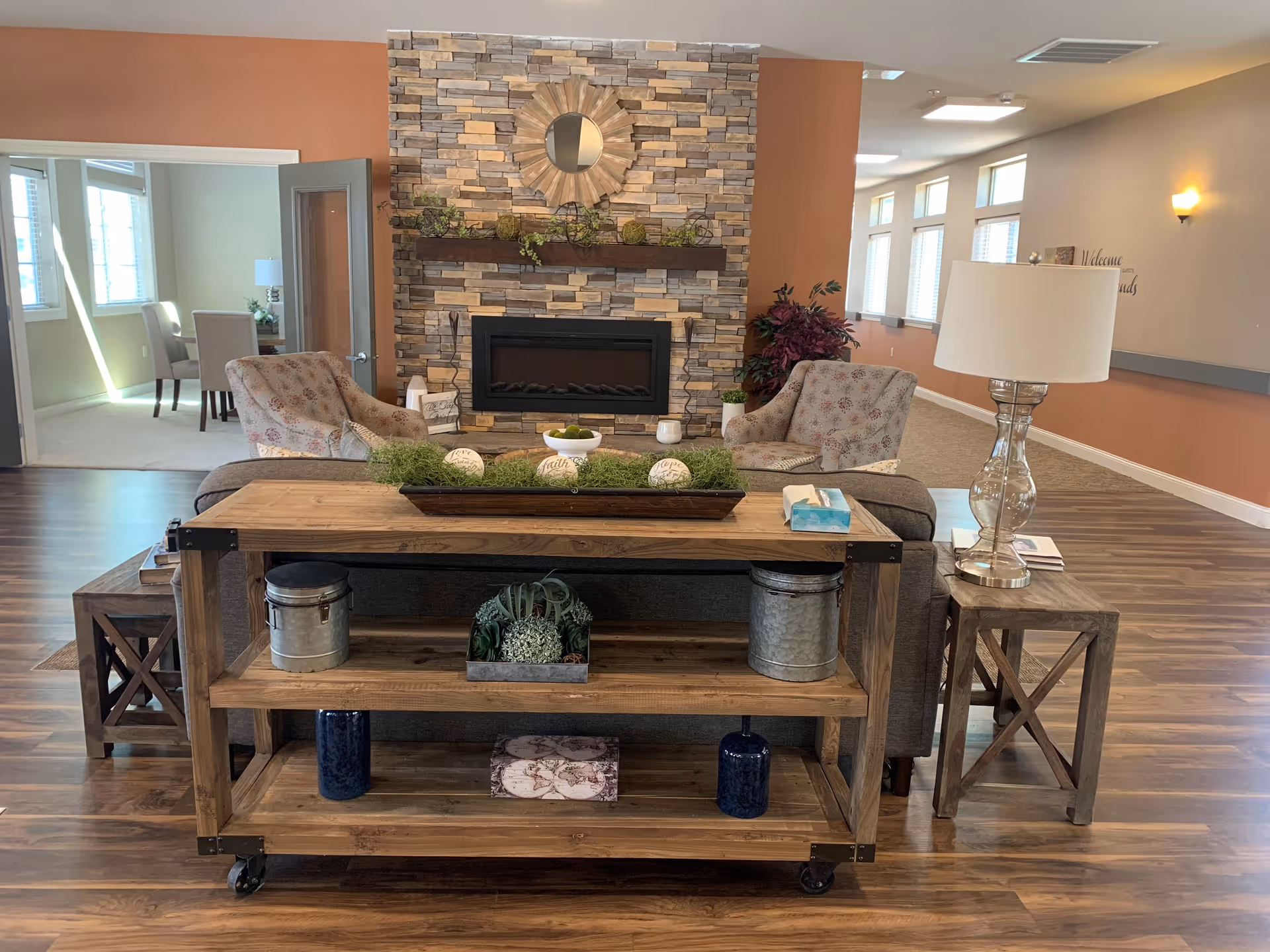 A cozy living room area with a stone fireplace as the focal point. In front of the fireplace are two patterned armchairs and a gray sofa. A wooden console table with decorative items such as plants and containers is placed behind the sofa. To the right of the sofa is a wooden side table with a glass lamp. The room has wood flooring and warm-toned walls, with a hallway and a dining area visible in the background.