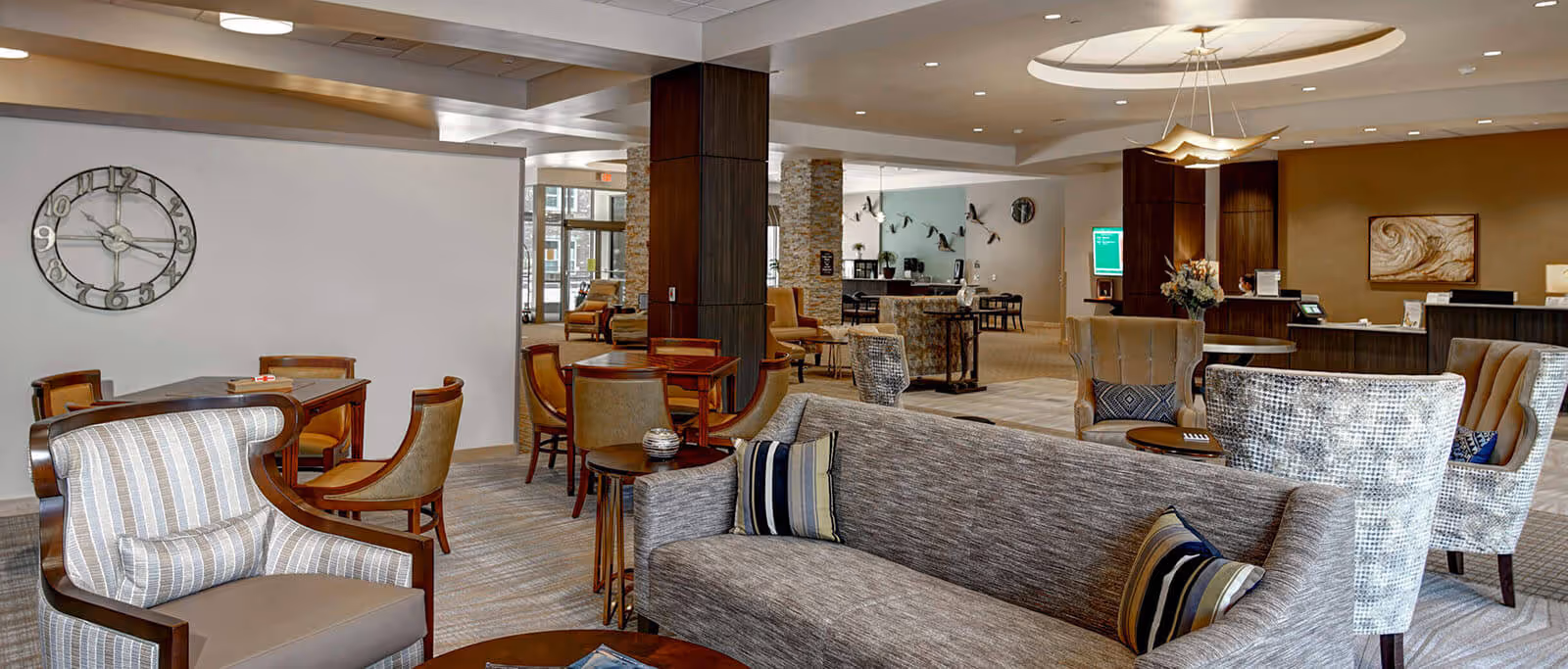 A spacious and well-lit senior living facility common area with comfortable seating including armchairs and sofas, wooden tables with chairs, a large wall clock, decorative wall art, and a reception desk in the background. The room features modern lighting fixtures and a neutral color palette with beige and brown tones.